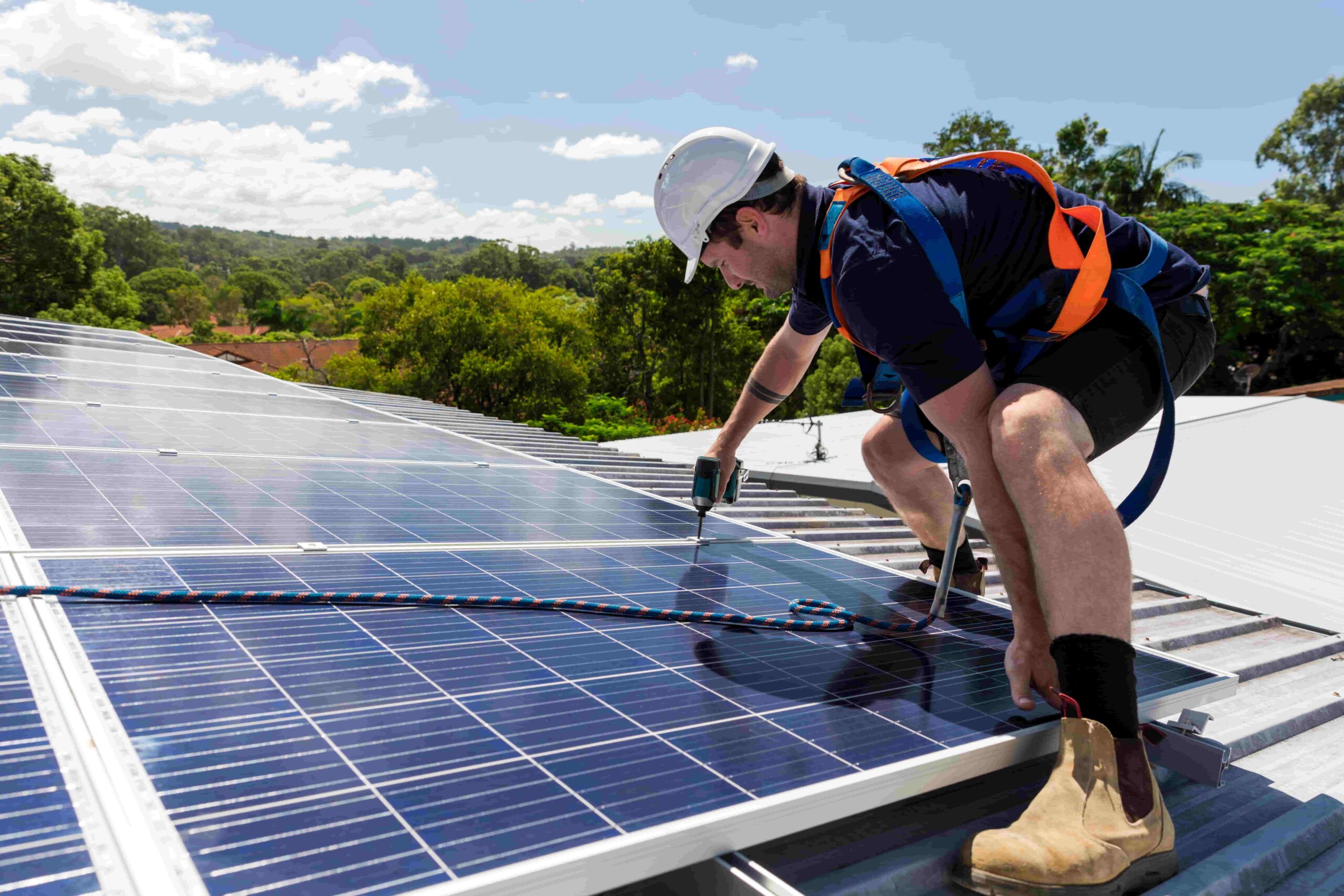 A worker in safety gear and a hard hat installs solar panels on a metal roof for DMG Solar using a power drill on a sunny day, with trees and blue sky in the background. Learn more about DMG Solar’s expert installation services. A worker in safety gear and a hard hat installs solar panels on a metal roof for DMG Solar using a power drill on a sunny day, with trees and blue sky in the background. Learn more about DMG Solar’s expert installation services.