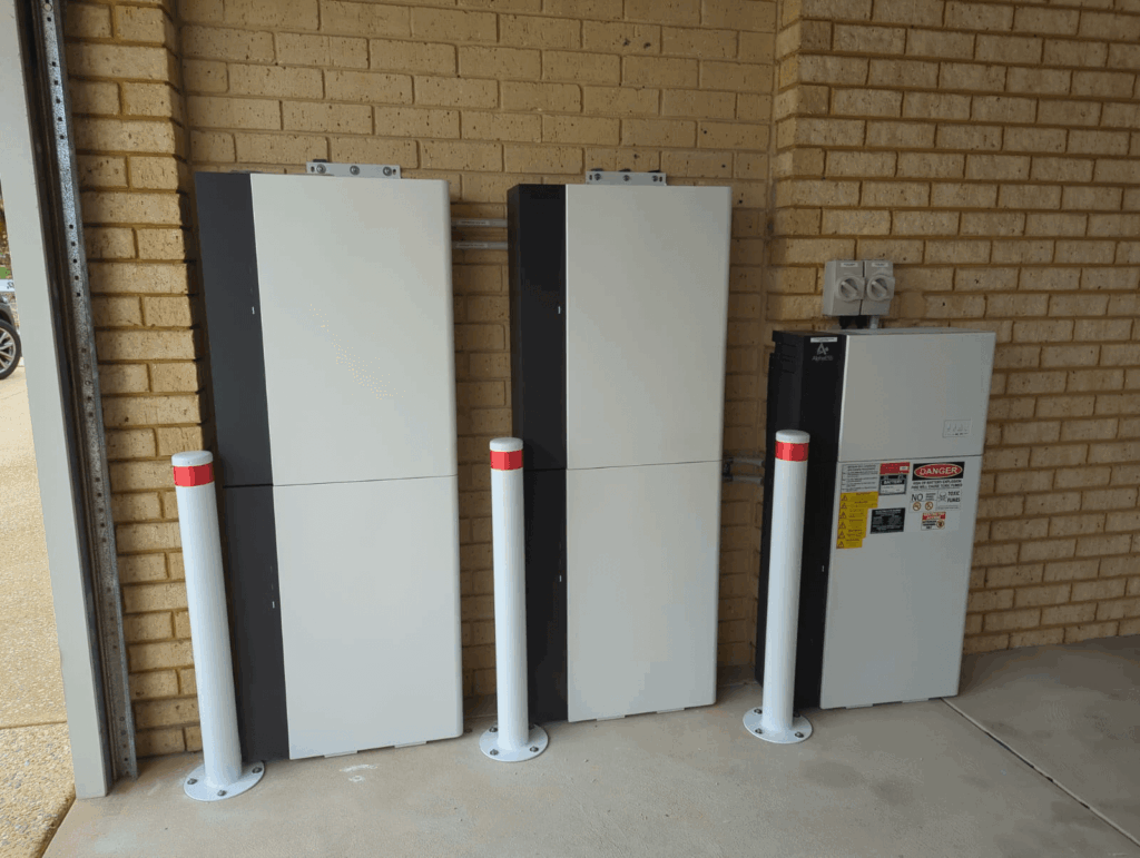 Three large 50 kWh DC-Coupled Energy Storage units and a control panel with warning labels are mounted on a brick wall indoors in Alexander Heights, with three white and red safety bollards in front of the units.