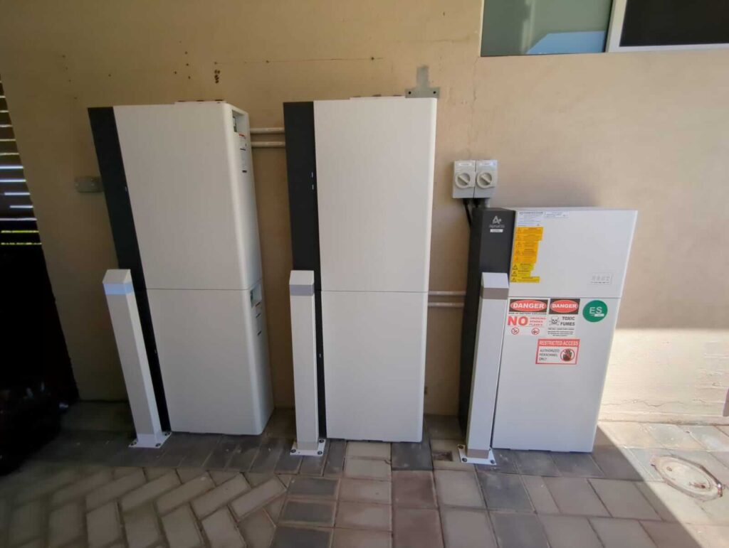 Three white battery storage units are installed side by side against a beige wall, complementing the tiled flooring below. The rightmost unit features safety labels and warning signs—part of a recent Ballajura solar installation.