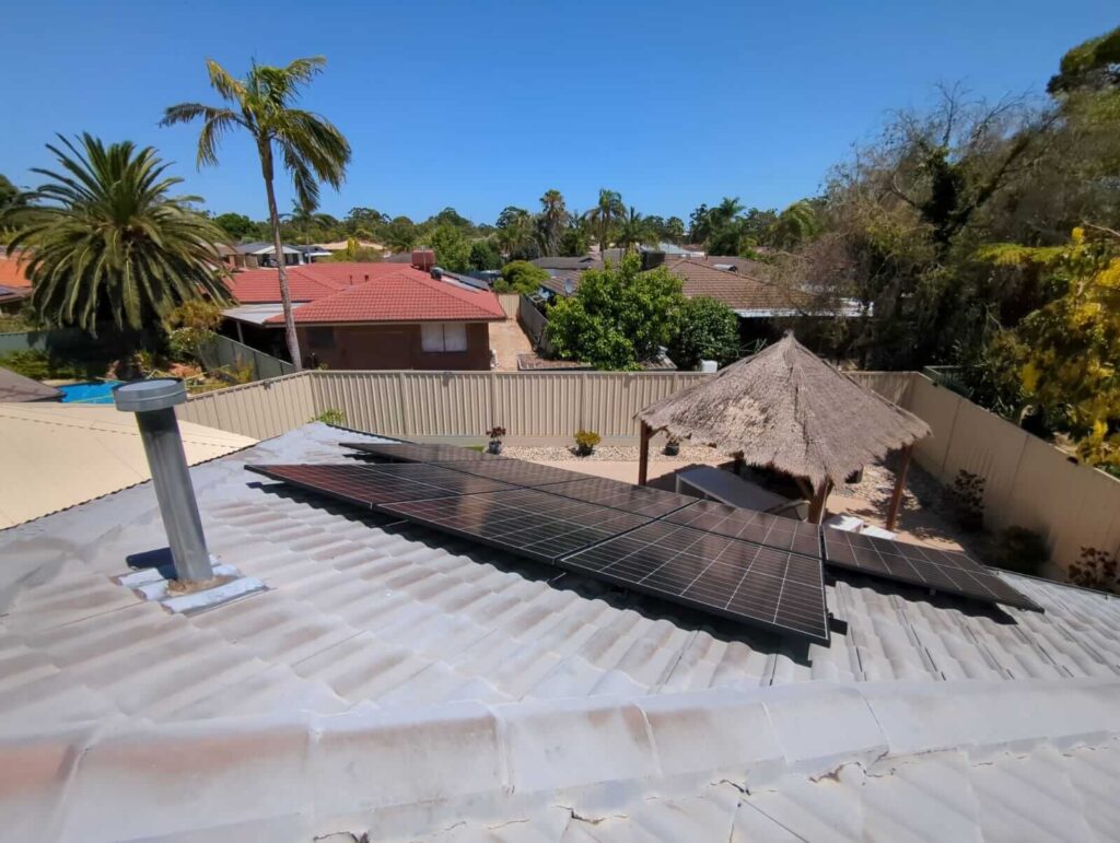 Ballajura solar installation featuring 9.97 kW solar panels on a corrugated roof, with lush greenery, a thatched gazebo, and neighbouring houses beneath a clear blue sky.