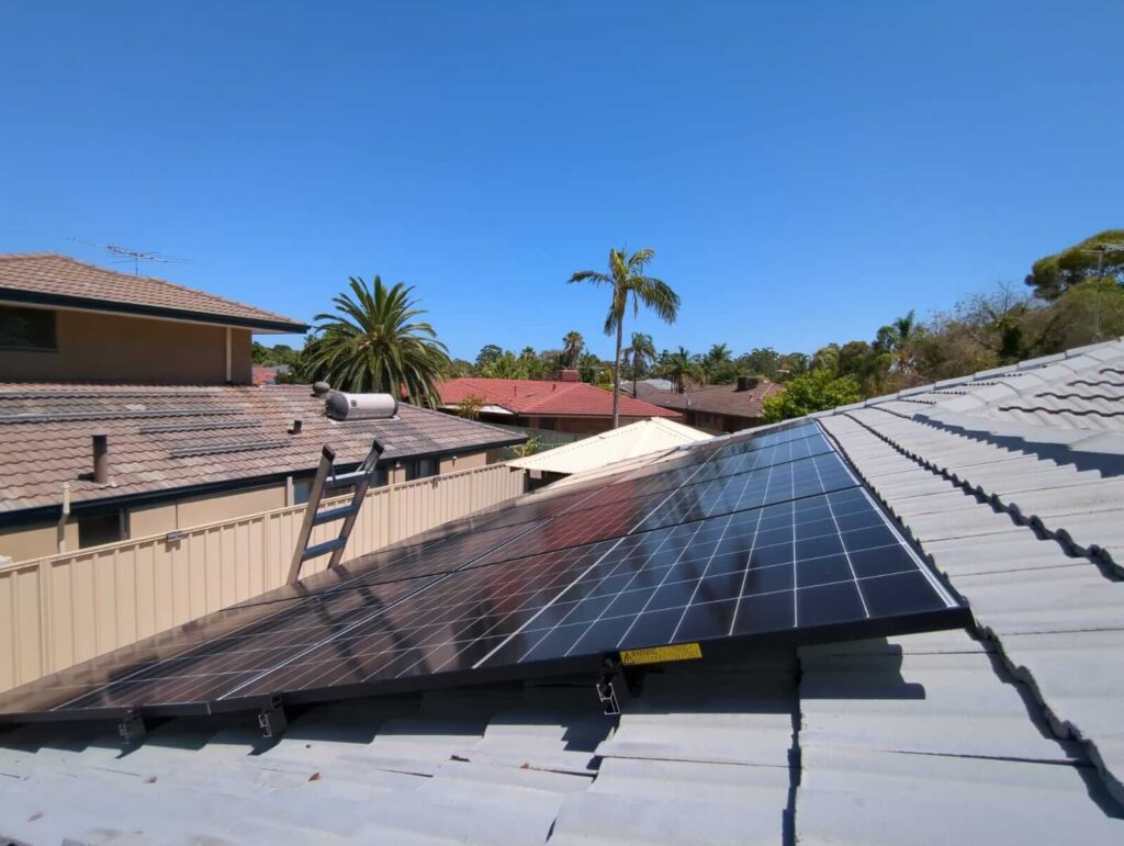A 9.97 kW solar system installed on a sloping rooftop under a clear blue sky in Ballajura, with neighbouring houses, palm trees, and a ladder visible in the background.