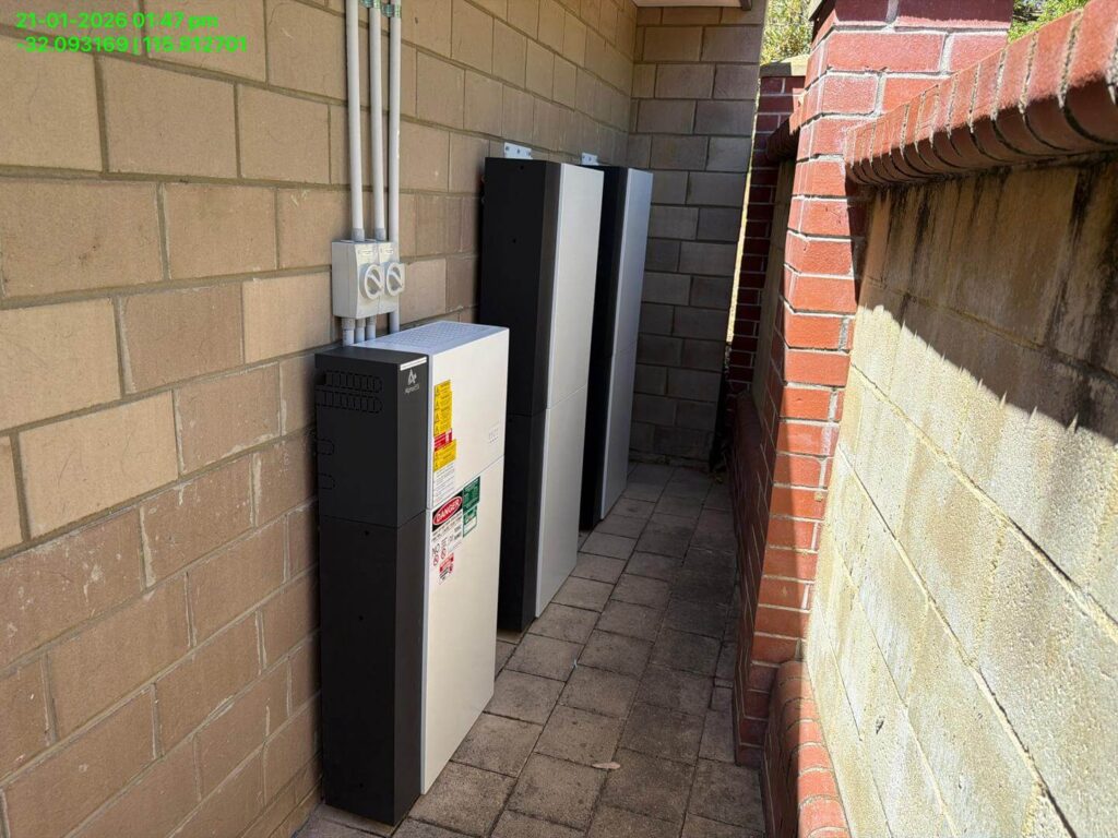 Three 9.975kW solar battery storage units are installed along a brick and block wall in a narrow Bibra Lake outdoor walkway with paved flooring. Electrical conduit and sockets are mounted above the 50.5kWh battery storage units.