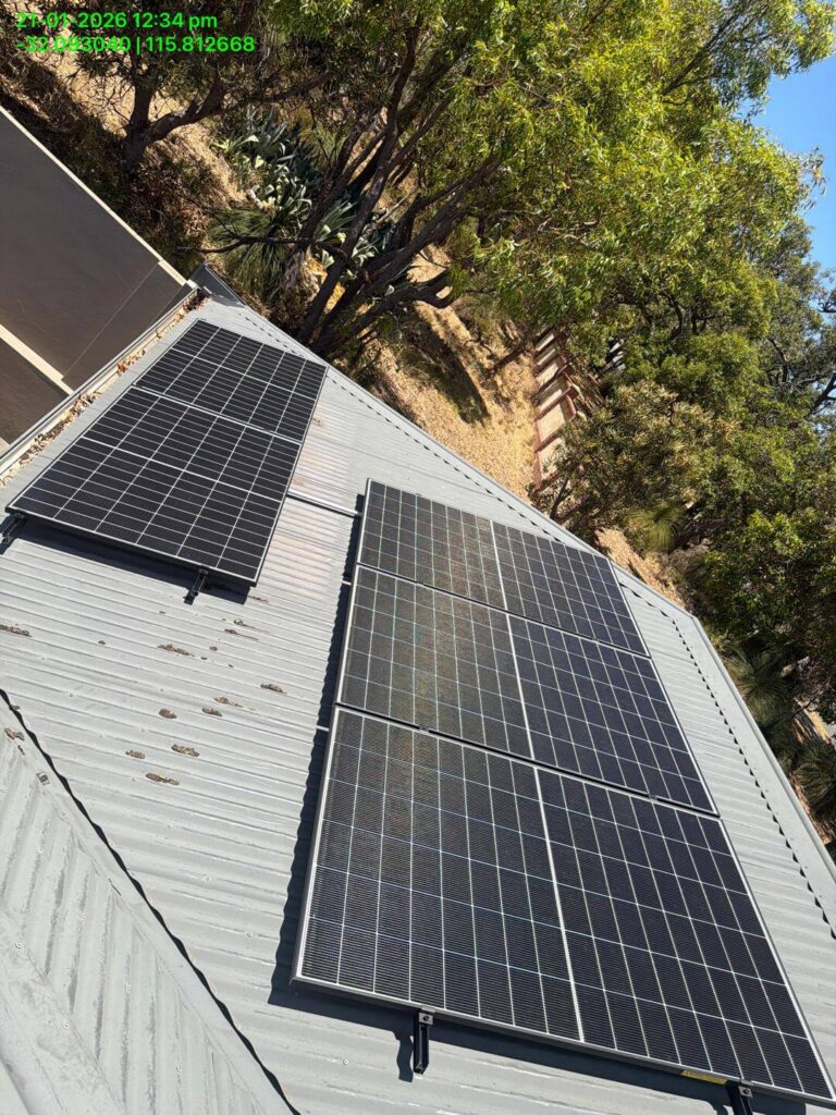 Three 9.975kW solar panels are mounted on the corrugated metal roof of a Bibra Lake building, surrounded by trees and dry vegetation. A timestamp and GPS coordinates appear in green text in the top left corner.