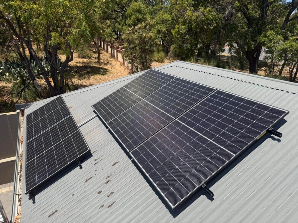 Solar panels are installed on the metal roof of a building in Bibra Lake, surrounded by trees and natural vegetation. The panels are positioned to capture sunlight efficiently, with potential for integration with battery storage systems.
