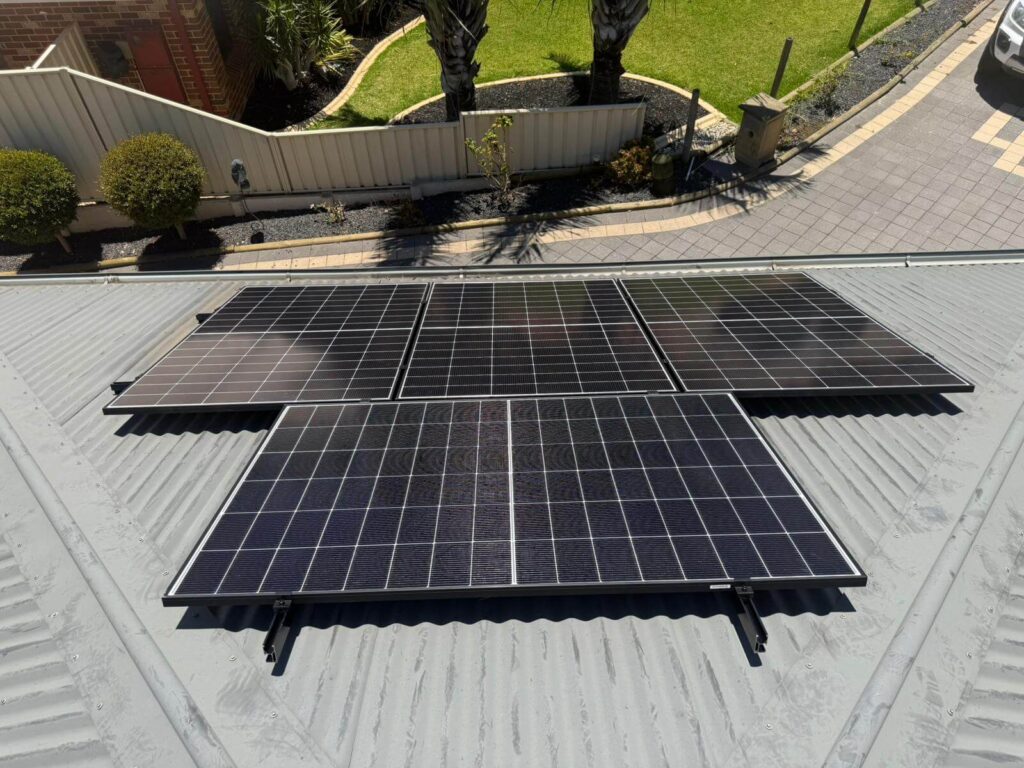 Four solar panels are installed on a corrugated metal rooftop in Bibra Lake. In the background, a neatly kept garden with trimmed bushes and a paved drive is visible, part of a 9.975kW solar system with 50.5kWh battery storage.
