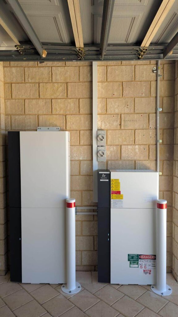 Two white electrical battery units, part of a 50.5kWh Battery Storage setup, are mounted on a brick wall in a garage and separated by metal conduit. Red-and-white safety bollards stand in front, with warning labels visible on the right unit.