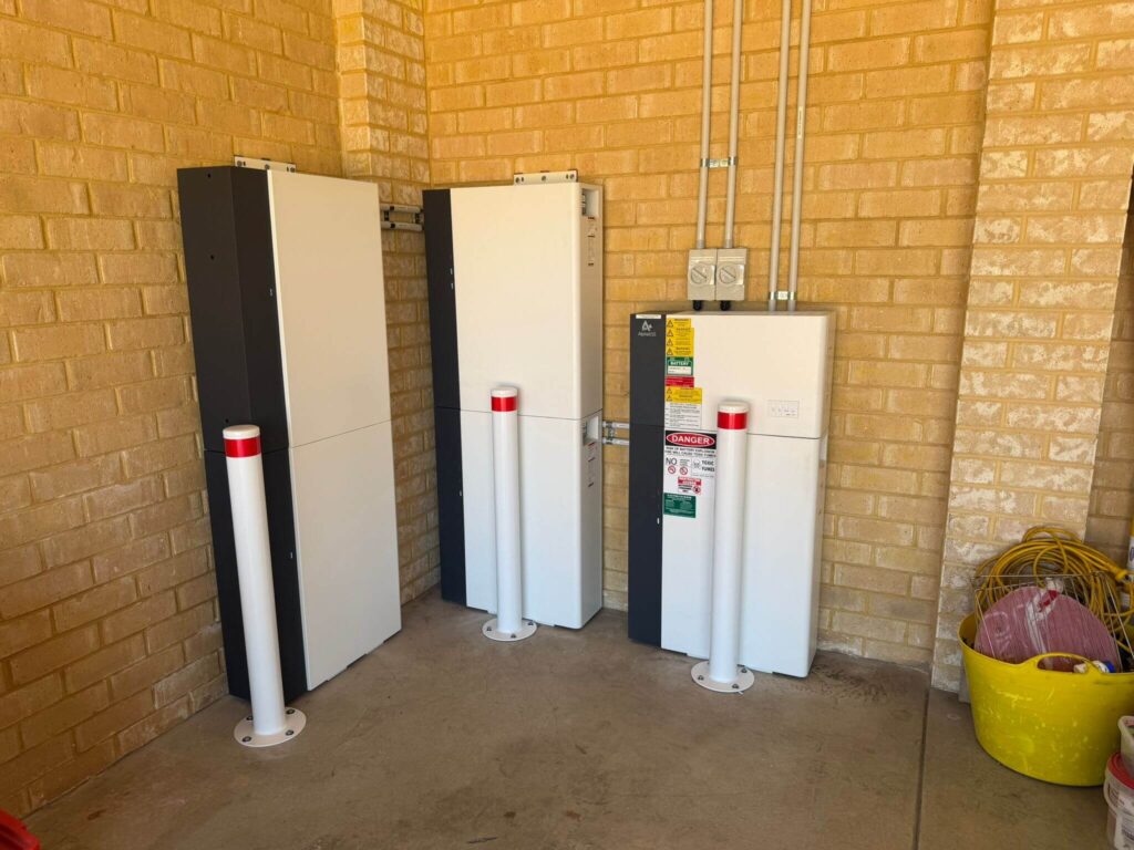 Three wall-mounted electrical units, part of a 50.5kWh battery storage system, with warning labels and protective white bollards are installed against a light brick wall in a clean utility area in Canning Vale. Electrical cables and a yellow bucket are visible nearby.