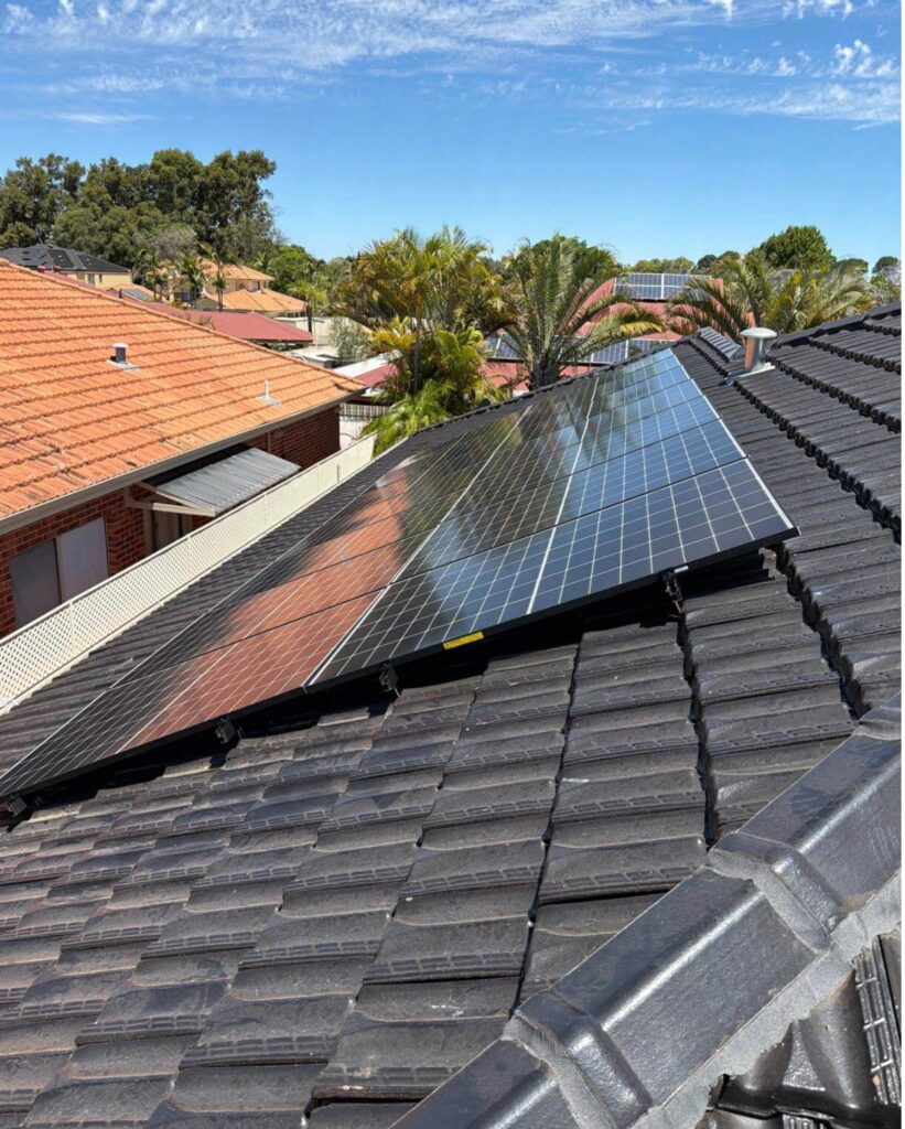 Solar panels installed on a dark tiled roof of a house in a suburban neighbourhood, featuring Battery Storage for enhanced efficiency. Red-tiled roofs, trees, and a partly cloudy blue sky complete this Canning Vale installation.