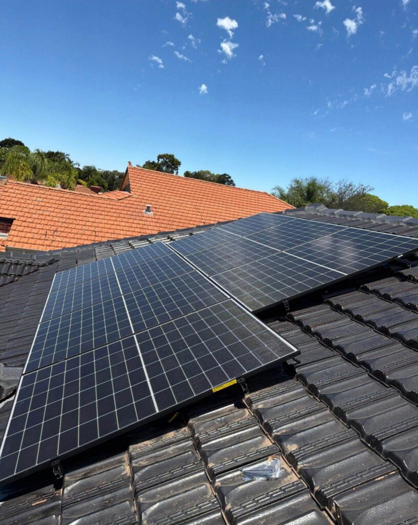 Rooftop solar panels from a Canning Vale solar installation gleam on a black-tiled roof under a blue sky with scattered clouds; red rooftops and green trees enhance the scenic backdrop.