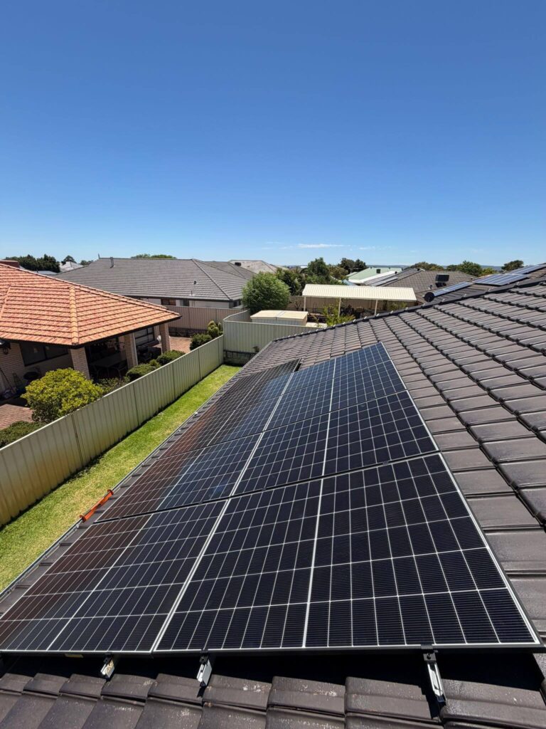 A row of 9.975kW solar panels is installed on the tiled roof of a Canning Vale home in a suburban neighbourhood, with neighbouring houses, fences, and a clear blue sky in the background.