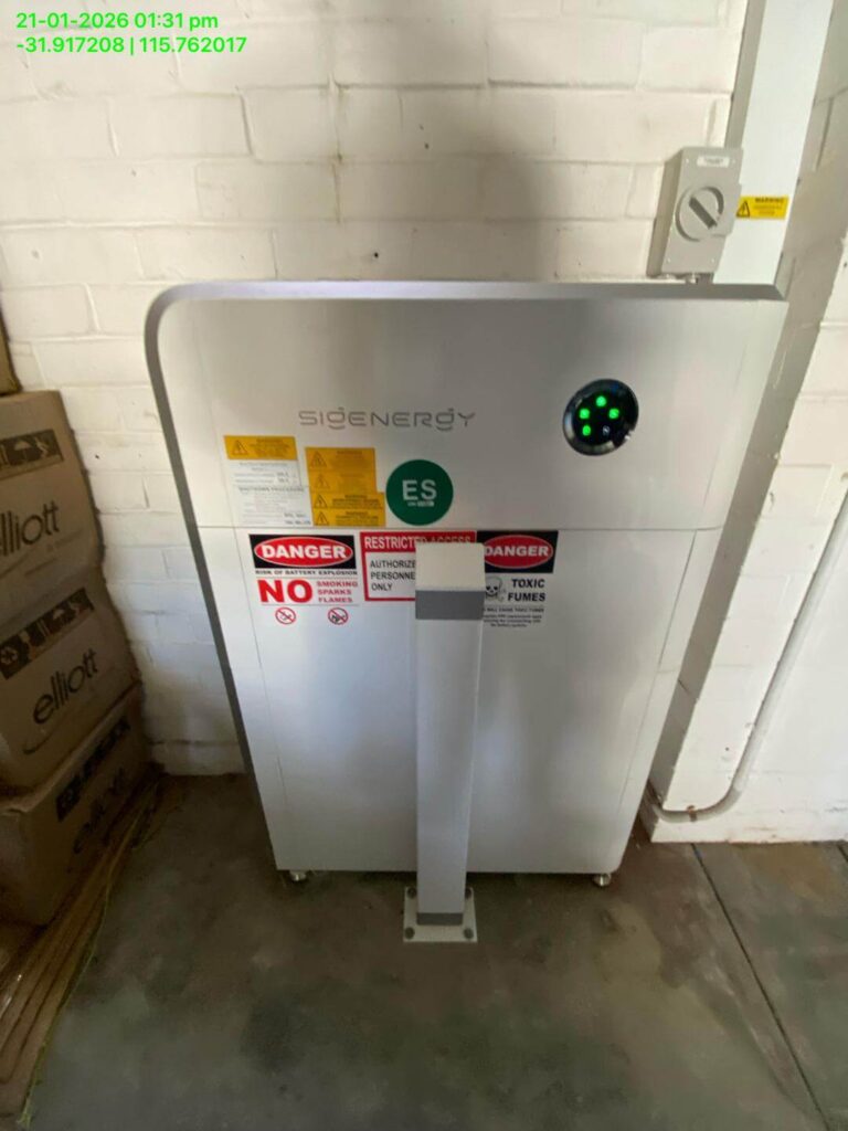 A white Sigenergy EC10TP battery unit with 24kWh storage and warning labels is installed against a white brick wall in a garage. Cardboard boxes are stacked on the left. Green coordinates and a timestamp appear at the top left.