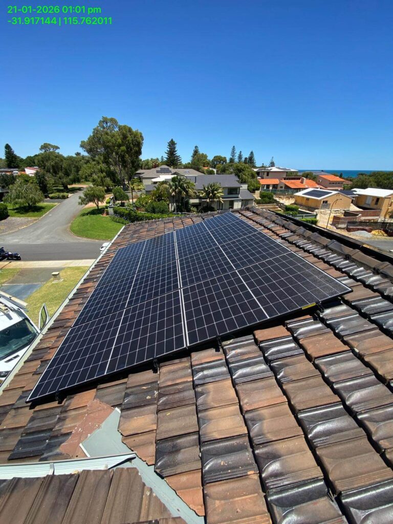 A 15.75kW solar array, paired with a Sigenergy EC10TP and 24kWh storage, is installed on a tiled rooftop in a suburban neighbourhood under clear skies, with trees, houses, and a glimpse of the sea in the background. Date, time, and coordinates appear at top left.