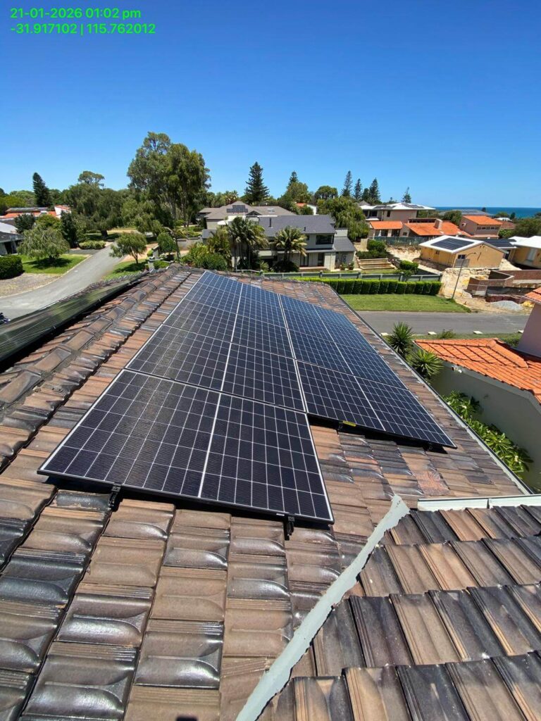 A row of solar panels, featuring a 15.75kW solar system and 24kWh storage, installed on a tiled residential rooftop with suburban houses, trees, and the sea visible in the background under a clear blue sky.