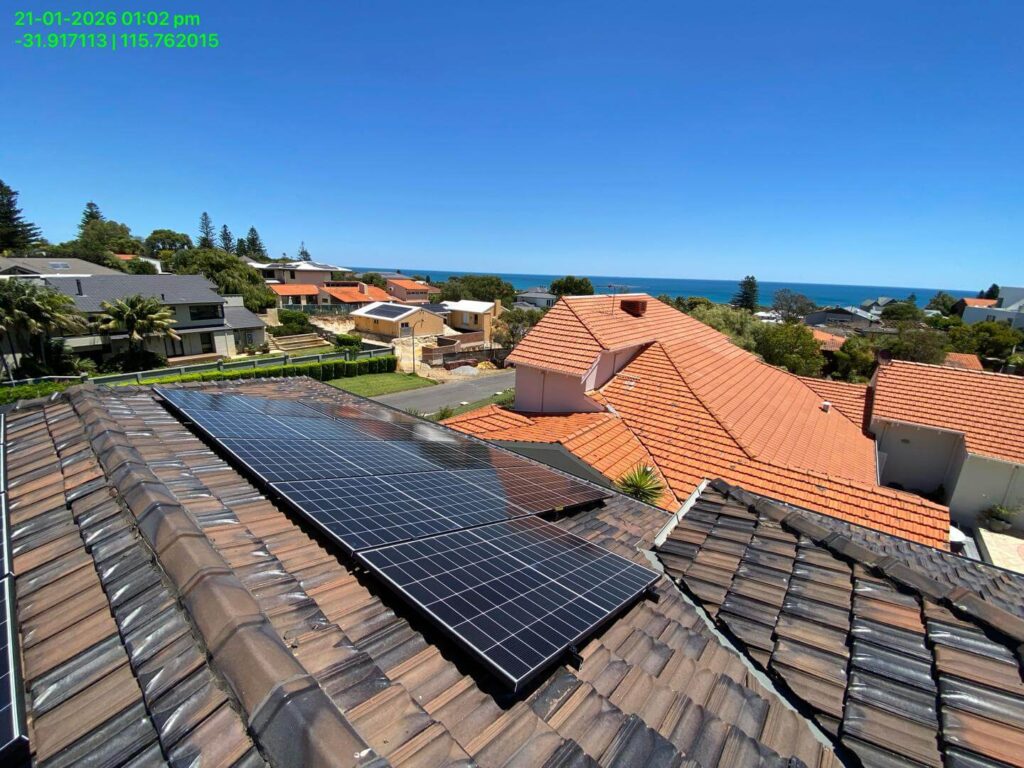 Solar installation featuring Sigenergy EC10TP panels on a tiled rooftop overlooks a suburban neighbourhood with orange roofs, green trees, and the sea under a clear blue sky. A 24kWh storage system ensures reliable energy day and night.