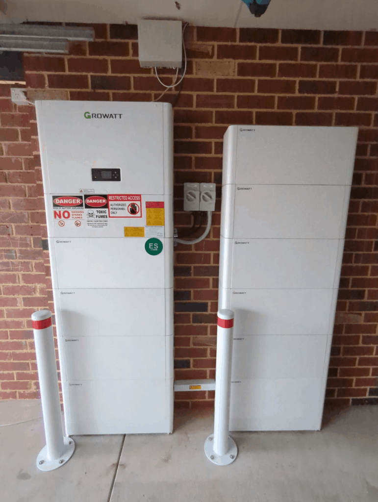 Two white Growatt energy storage units are mounted on a brick wall in Ellenbrook, with visible warning labels and electrical panels. Two white safety bollards with red stripes are installed in front of the units.