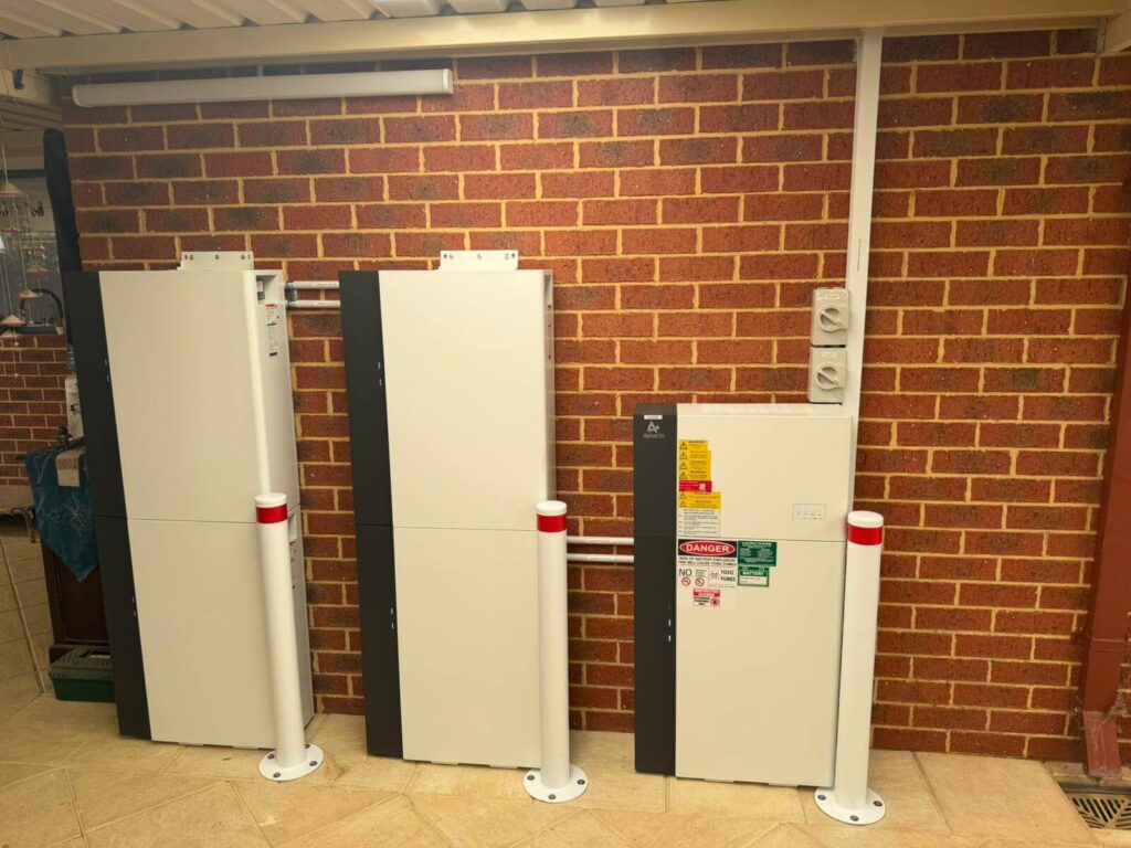 Three white battery storage units with black vertical stripes are mounted against a red brick wall, part of a 9.975 kW solar inverter installation. Warning labels and protective bollards stand in front, with electrical conduits running above and beside the units.