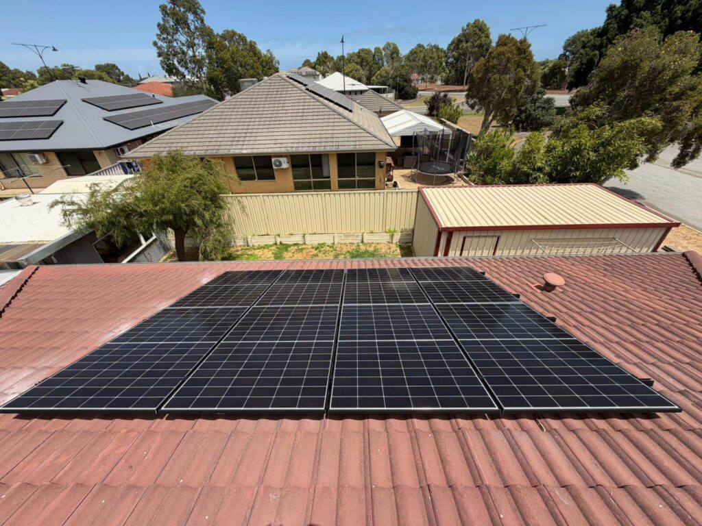 Rows of solar panels and an inverter installed on a red-tiled house roof, with neighbouring houses, trees, and a clear blue sky in the background.