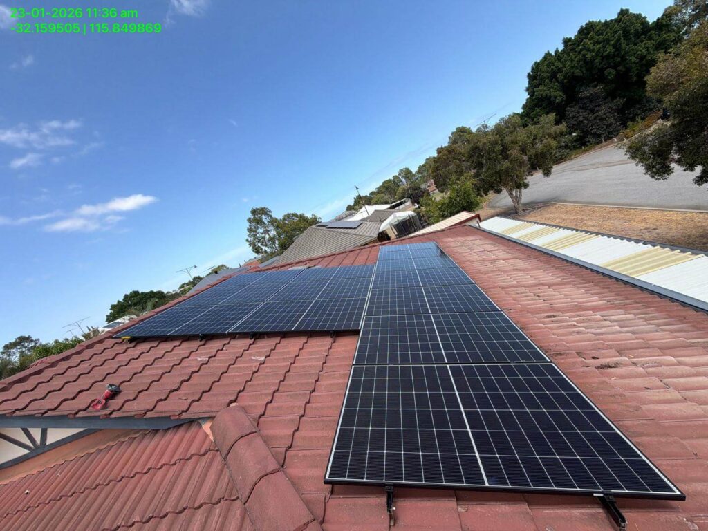 Solar installation featuring panels on a red-tiled rooftop under a clear sky, surrounded by trees and neighbouring houses. A timestamp and GPS coordinates are visible in green text at the top left corner of the image.