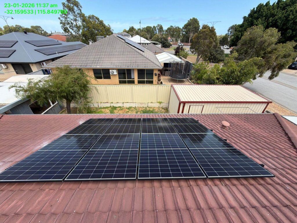 Rooftop solar panels paired with an inverter are installed on a reddish-brown tiled roof, overlooking neighbouring houses, a shed, trees, and a suburban street under a clear sky. Date and coordinates appear in green text.