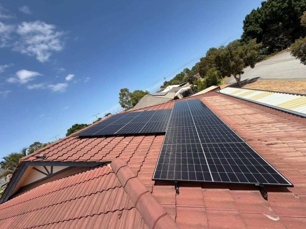 Solar installation on the red-tiled roof of a Hammond Park home, with trees and other buildings visible in the background beneath a clear blue sky.