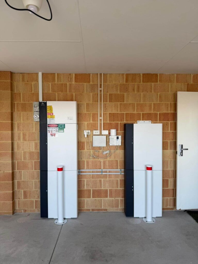 Two white 40 kWh battery storage units with warning labels are mounted against a brick wall in Harrisdale. Electrical panels and sockets are between them, with two white safety bollards with red tops in front of the units.