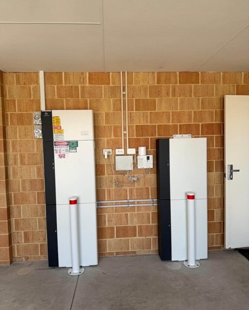 Two tall white battery units with control panels, part of a 40 kWh battery storage system, are installed against a brick wall indoors, with pipes and electrical boxes above and between them. Two white safety bollards stand in front, and a closed door is visible on the right.