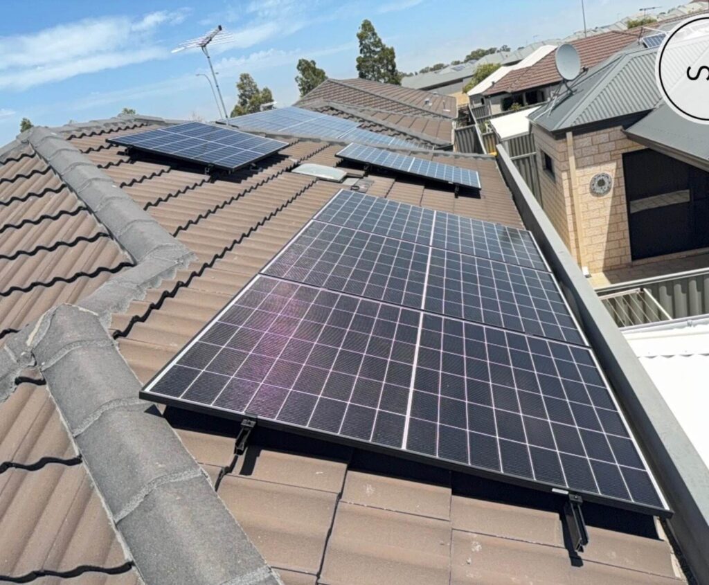 Solar panels with a 7.125 kW solar system are mounted on a brown tiled house roof under a blue sky, with neighbouring houses in the background. The panels are angled efficiently, and an inverter is installed for optimal energy use.