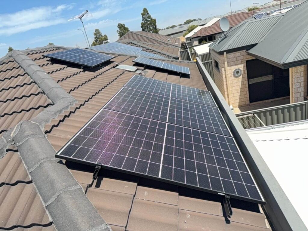 Solar panels, part of a 7.125 kW solar system in Harrisdale, are installed on a tiled residential roof. Additional panels are visible on neighbouring homes, with trees and satellite dishes set against a clear sky.