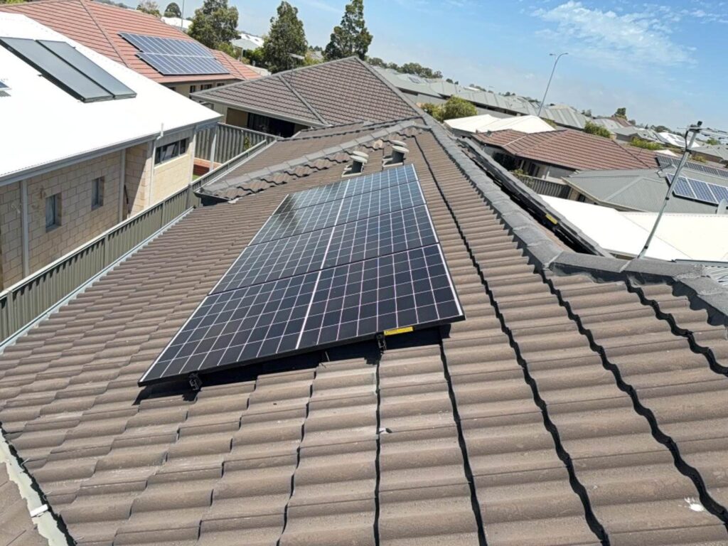 Solar panels installed on the brown tiled roof of a Harrisdale home in a suburban neighbourhood, featuring a 7.125 kW solar system with 40 kWh battery storage; nearby houses, some with panels too, under a clear blue sky.