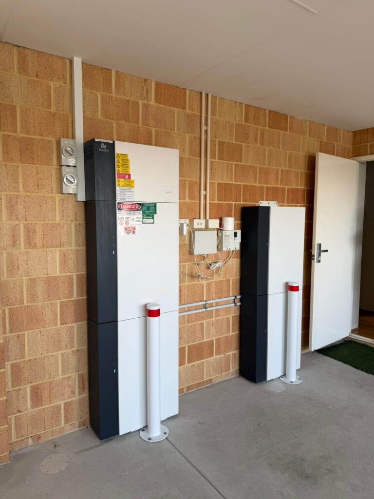Two tall, rectangular 40 kWh battery storage units are mounted on a brick wall in a Harrisdale garage. The units display warning labels, have adjacent power switches, protective white bollards in front, and an open door to the right.