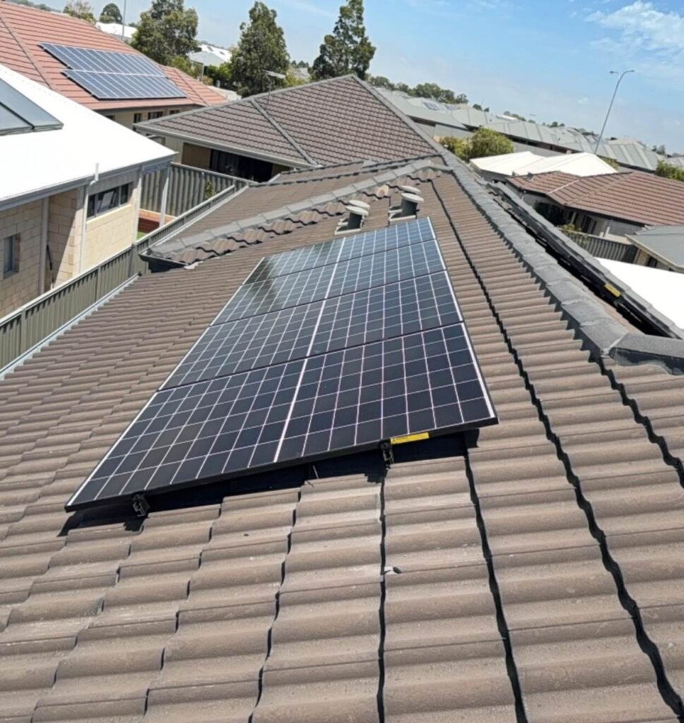 Two rows of solar panels, part of a 7.125 kW solar system, are installed on the tiled roof of a house in a suburban neighbourhood. Other houses with similar roofs and some with solar panels can be seen in the background.