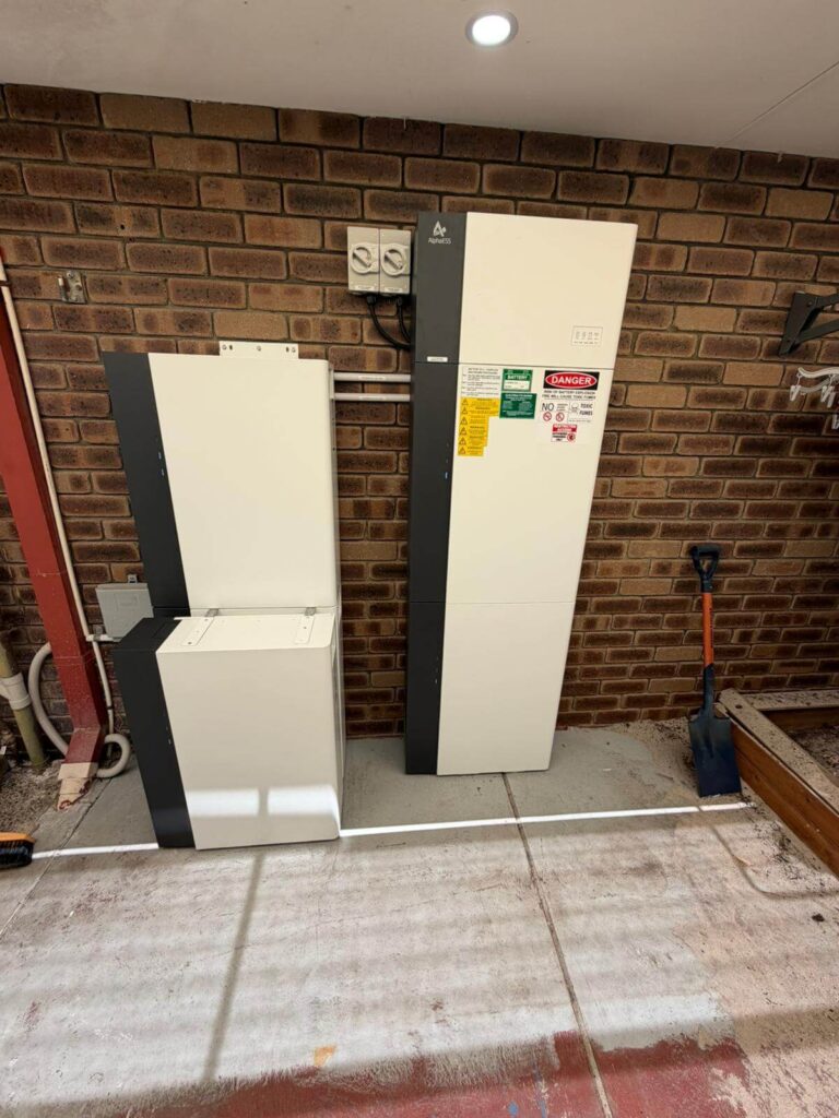 Two large 50.5kWh battery storage units are installed against a brick wall in a covered outdoor area at Parkwood. The units have labels and warning stickers, whilst a blue spade leans against the wall nearby on the right.