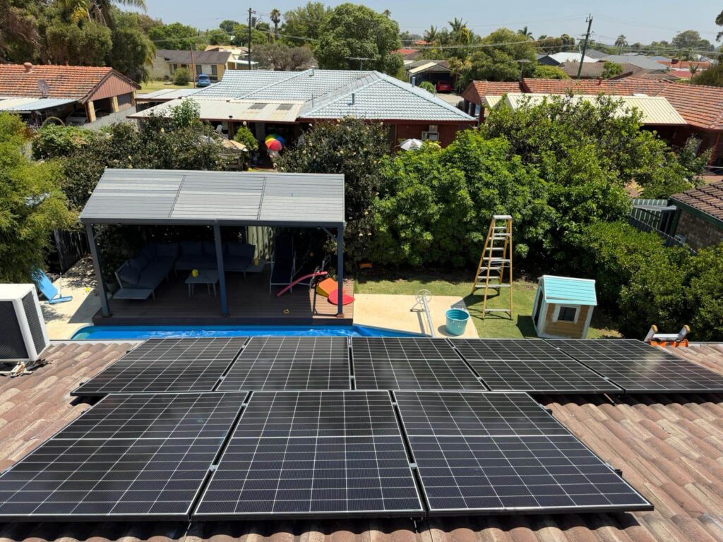 9.975kW solar panels are installed on a tiled roof in Parkwood, overlooking a back garden with a small pool, covered outdoor seating, ladder, playhouse, lush trees, and neighbouring houses; 50.5kWh battery storage is included.