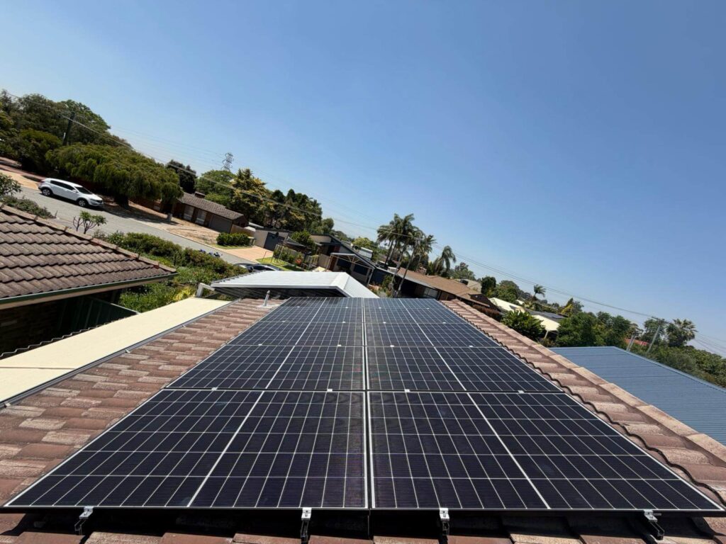 Rows of 9.975kW solar panels installed on a tiled Parkwood rooftop, with a clear blue sky above and suburban houses, trees, and parked cars visible in the background.