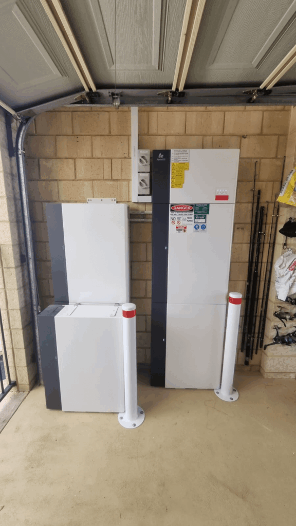 Two large, white battery storage units with warning labels are installed against a brick wall in a Southern River garage. Two white protective bollards are positioned in front of the units for safety.