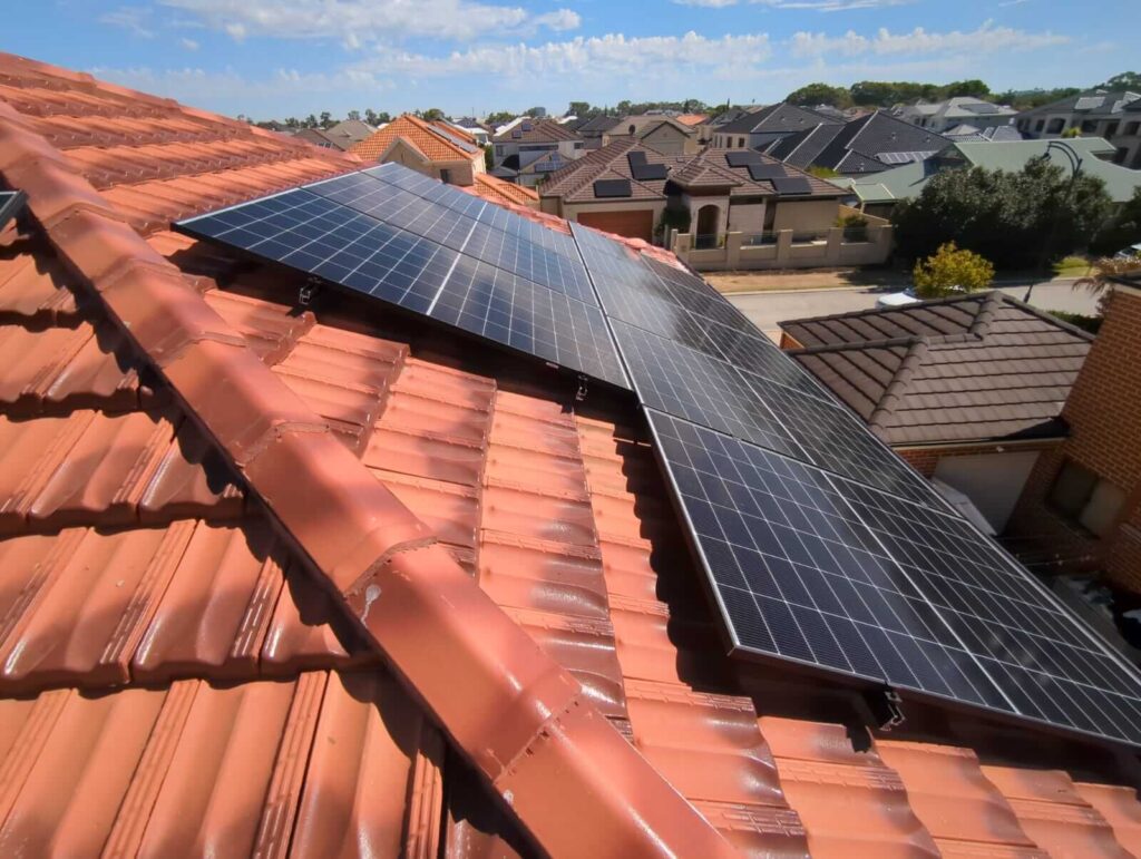 Rows of 11 kW solar panels are installed on the red-tiled roof of a house in a suburban neighbourhood, with multiple houses and trees visible in the background under a partly cloudy sky. Growatt battery storage supports efficient energy use.