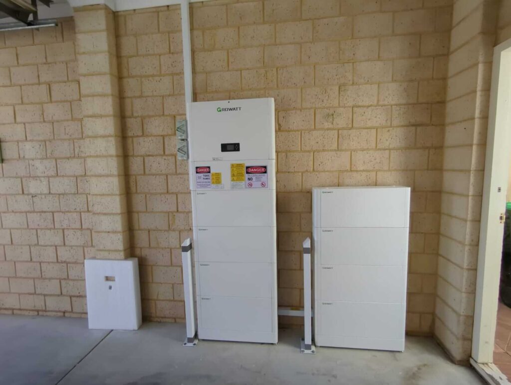 Large white Growatt Battery Storage units are installed against a beige brick wall in a garage or utility area in Stirling. Safety warning labels are visible on the central unit, supporting an 11 kW solar system.