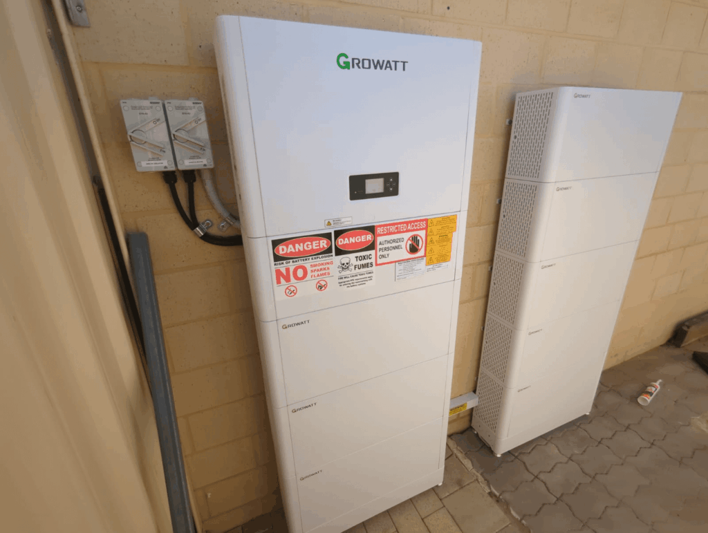 A Growatt AC-Coupled Storage System is mounted on an outdoor brick wall, with warning and safety labels visible. Another similar battery unit, part of a Swan View installation with 40 kWh storage, stands nearby on a tiled ground.