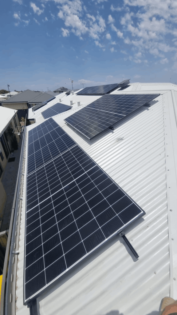 Rows of 9.97 kW solar panels installed on a white corrugated metal rooftop in Treeby under a partly cloudy blue sky, complemented by 50 kWh battery storage, with neighbouring buildings visible in the background.