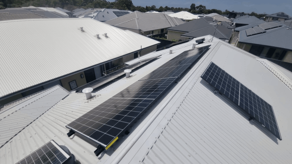 Rows of 9.97 kW solar panels are installed on the white metal roof of a house in Treeby, a suburban neighbourhood where other homes also showcase solar arrays and some feature 50 kWh battery storage systems in the background.