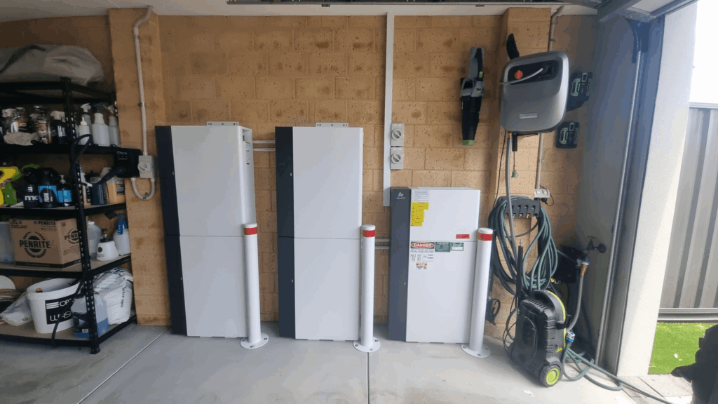 Three large 50 kWh battery storage units and an electrical box are mounted on a Treeby garage wall. Nearby are shelves with various items, a wall-mounted hose reel, and a pressure washer on the floor. The garage door is partially open.