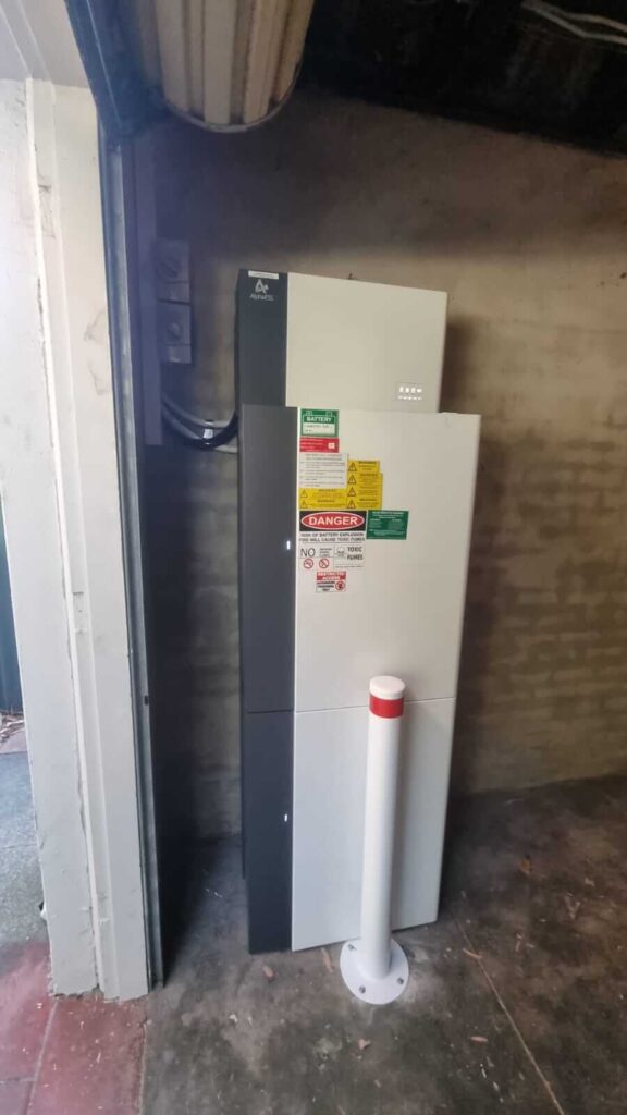 A large 40kWh battery storage unit with multiple warning and safety labels is installed against a brick wall in a Wembley garage. A white bollard is positioned in front of the unit for protection.