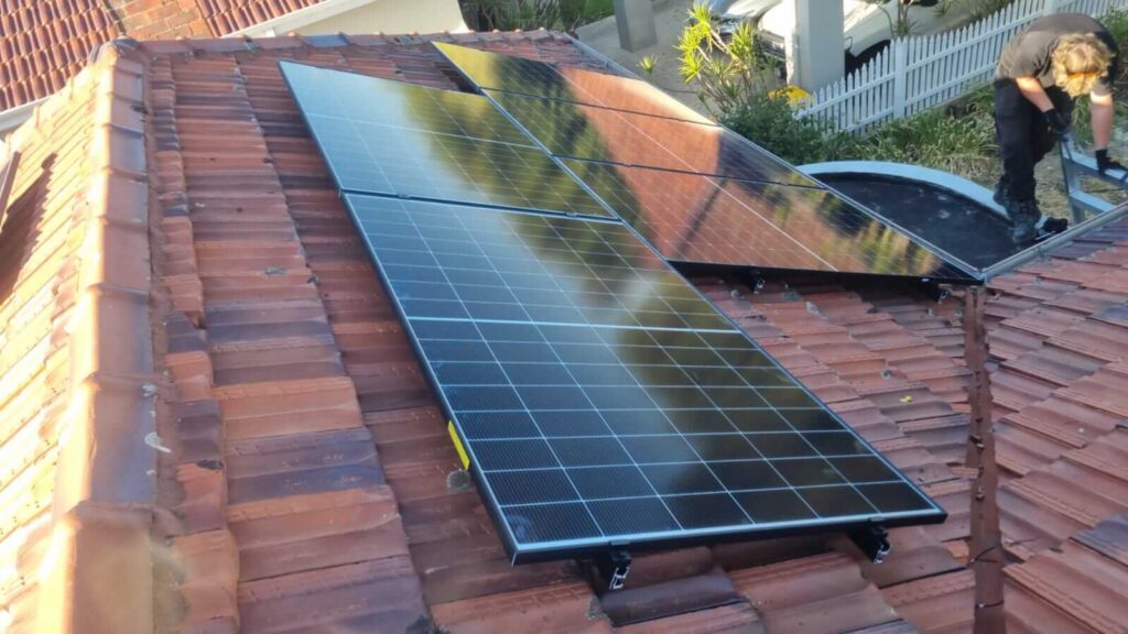 Several black solar panels, part of a 9.5kW solar system, are installed on a red-tiled sloped roof in Wembley, with a person working nearby. A white fence and plants are visible in the background.