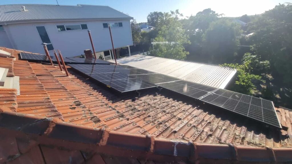 Several solar panels are mounted on a sloped, red-tiled roof in Wembley, paired with 9.5kW solar and 40kWh battery storage. A neighbouring building with a metal roof and trees are visible in the background on a sunny day.