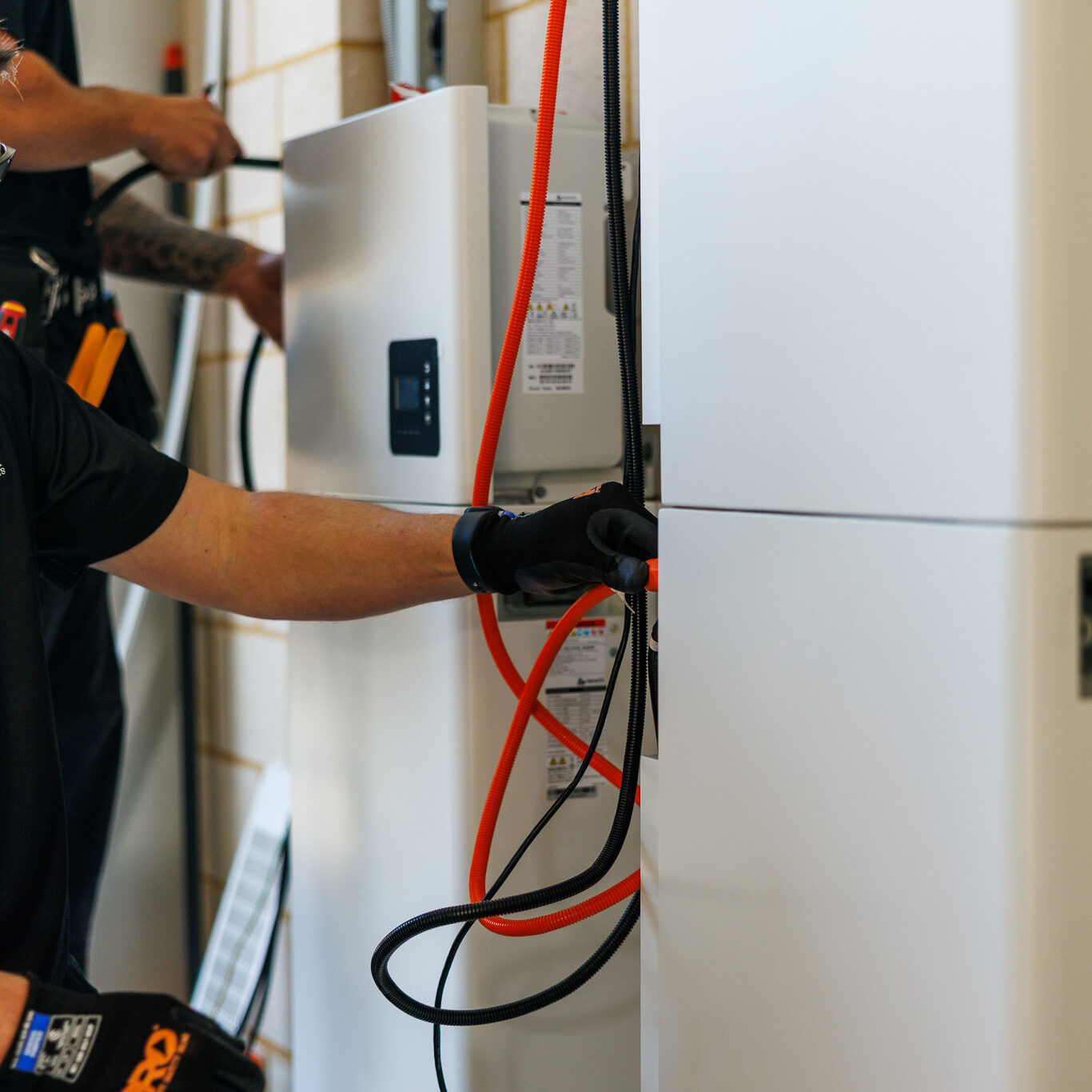 A technician wearing gloves and sunglasses connects orange cables to a white electrical unit during a Home Battery Installation, with another person working in the background.