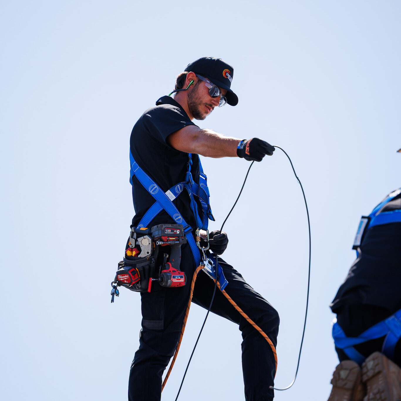 A construction worker wearing safety gear and a harness stands on a sloped surface, handling an electrical wire as part of repairs against a clear blue sky.